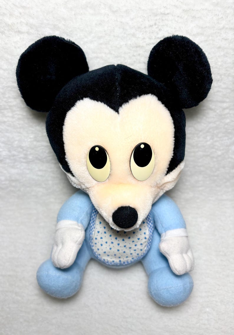 May include: A soft toy Mickey Mouse with black ears and head. The doll has a white face and a blue outfit with a bib featuring a white heart pattern. The doll's eyes are large and black.