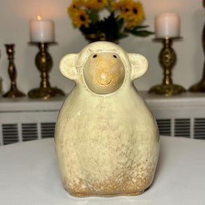 Puede incluir: Una figura de oveja de cerámica con un esmalte de color crema y un acabado texturizado de color marrón moteado en la base. La oveja tiene una cabeza redondeada, orejas prominentes y una cara sencilla y lisa con dos pequeños puntos oscuros como ojos.