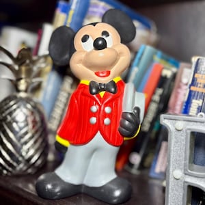 Peut inclure: Figurine en c&eacute;ramique de Mickey Mouse portant une veste rouge, un pantalon gris et des chaussures noires. La figurine tient un livre gris et porte un n&oelig;ud papillon noir. L'arri&egrave;re-plan comprend une d&eacute;coration d'ananas et une &eacute;tag&egrave;re de livres.