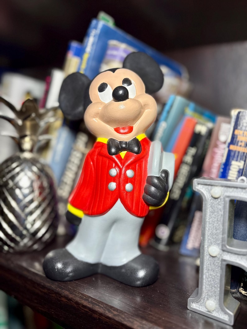 Peut inclure: Figurine en c&eacute;ramique de Mickey Mouse portant une veste rouge, un pantalon gris et des chaussures noires. La figurine tient un livre gris et porte un n&oelig;ud papillon noir. L'arri&egrave;re-plan comprend une d&eacute;coration d'ananas et une &eacute;tag&egrave;re de livres.