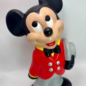 Peut inclure: Figurine en c&eacute;ramique de Mickey Mouse portant une veste rouge, un pantalon gris et des chaussures noires. La figurine a des oreilles noires, un n&oelig;ud papillon noir et tient un chapeau gris. La figurine a une expression souriante.