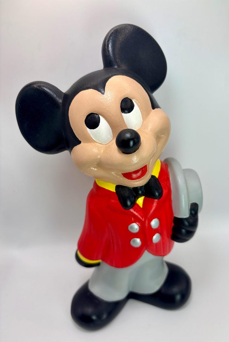 Peut inclure: Figurine en c&eacute;ramique de Mickey Mouse portant une veste rouge, un pantalon gris et des chaussures noires. La figurine a des oreilles noires, un n&oelig;ud papillon noir et tient un chapeau gris. La figurine a une expression souriante.
