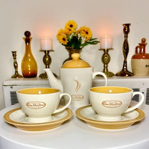 May include: A ceramic tea set featuring a teapot and two cups with matching saucers. The pieces are cream-coloured with tan accents and rims. Each item displays the Tim Hortons logo and the text Always Fresh, with a stylised steam graphic on the teapot.