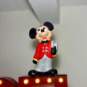Peut inclure: Figurine de Mickey Mouse portant une veste rouge &agrave; bordures jaunes, un pantalon gris et des chaussures noires. La figurine tient un objet blanc et porte un n&oelig;ud papillon noir. Elle est pos&eacute;e sur une lettre de chapiteau rouge.