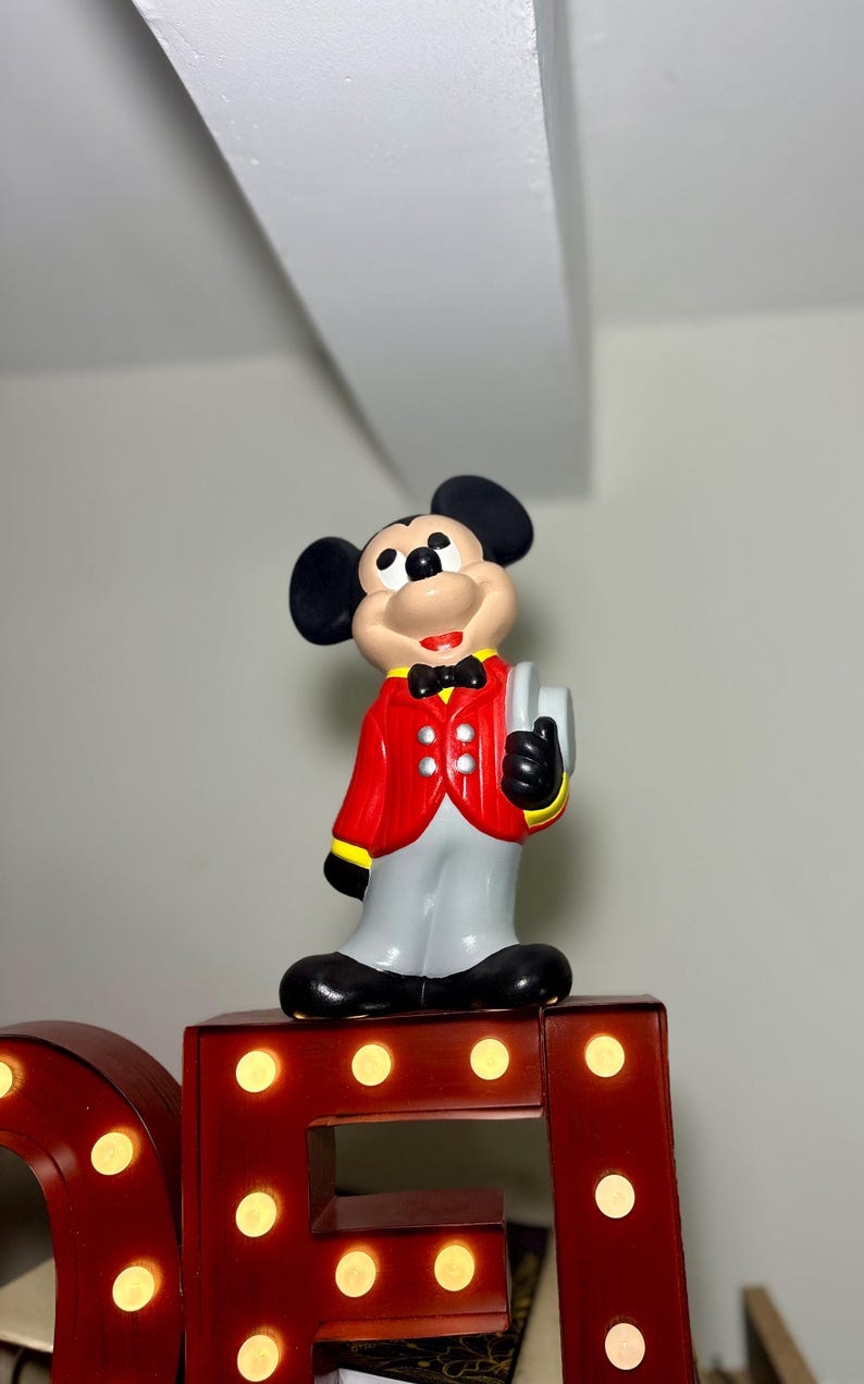 Peut inclure: Figurine de Mickey Mouse portant une veste rouge &agrave; bordures jaunes, un pantalon gris et des chaussures noires. La figurine tient un objet blanc et porte un n&oelig;ud papillon noir. Elle est pos&eacute;e sur une lettre de chapiteau rouge.