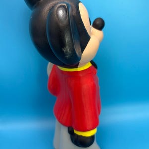 Peut inclure: Figurine en c&eacute;ramique de Mickey Mouse, vue de c&ocirc;t&eacute;. Mickey porte une veste rouge avec des bordures jaunes, un pantalon gris et des chaussures noires. Ses oreilles sont noires et son visage beige avec un nez noir. Le fond est bleu uni.