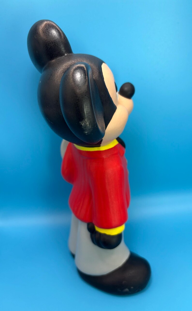 Peut inclure: Figurine en c&eacute;ramique de Mickey Mouse, vue de c&ocirc;t&eacute;. Mickey porte une veste rouge avec des bordures jaunes, un pantalon gris et des chaussures noires. Ses oreilles sont noires et son visage beige avec un nez noir. Le fond est bleu uni.