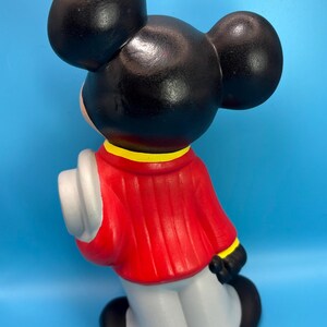 Peut inclure: Figurine en c&eacute;ramique d'un personnage de dessin anim&eacute; de souris, vue de dos. La figurine a de grandes oreilles noires, une veste rouge avec des bordures jaunes, des gants blancs et un pantalon gris. Il tient un chapeau blanc. Le fond est bleu clair uni.