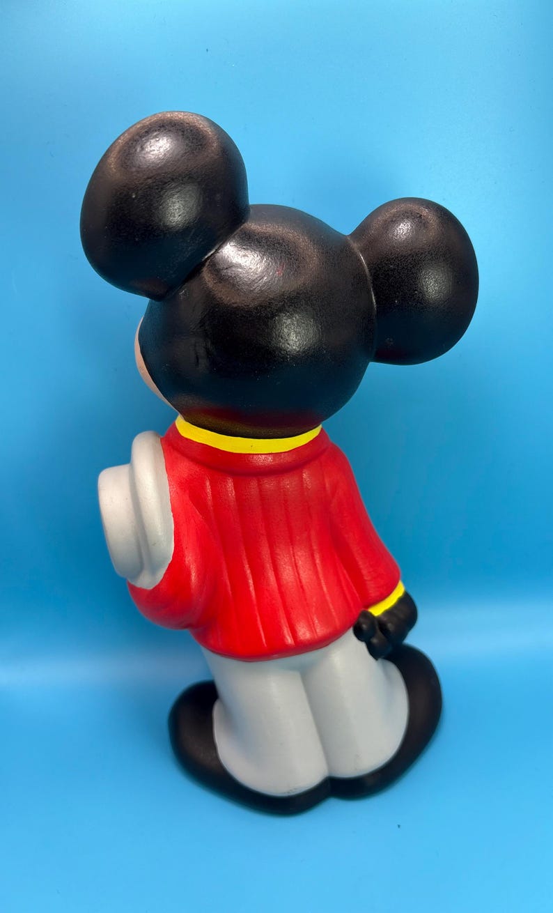 Peut inclure: Figurine en c&eacute;ramique d'un personnage de dessin anim&eacute; de souris, vue de dos. La figurine a de grandes oreilles noires, une veste rouge avec des bordures jaunes, des gants blancs et un pantalon gris. Il tient un chapeau blanc. Le fond est bleu clair uni.