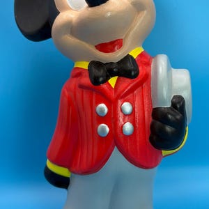 Peut inclure: Figurine en c&eacute;ramique de Mickey Mouse, v&ecirc;tu d'une veste rouge, d'un pantalon gris et de chaussures noires. La figurine porte un n&oelig;ud papillon noir, des oreilles noires et tient un objet blanc. Le fond est bleu uni.