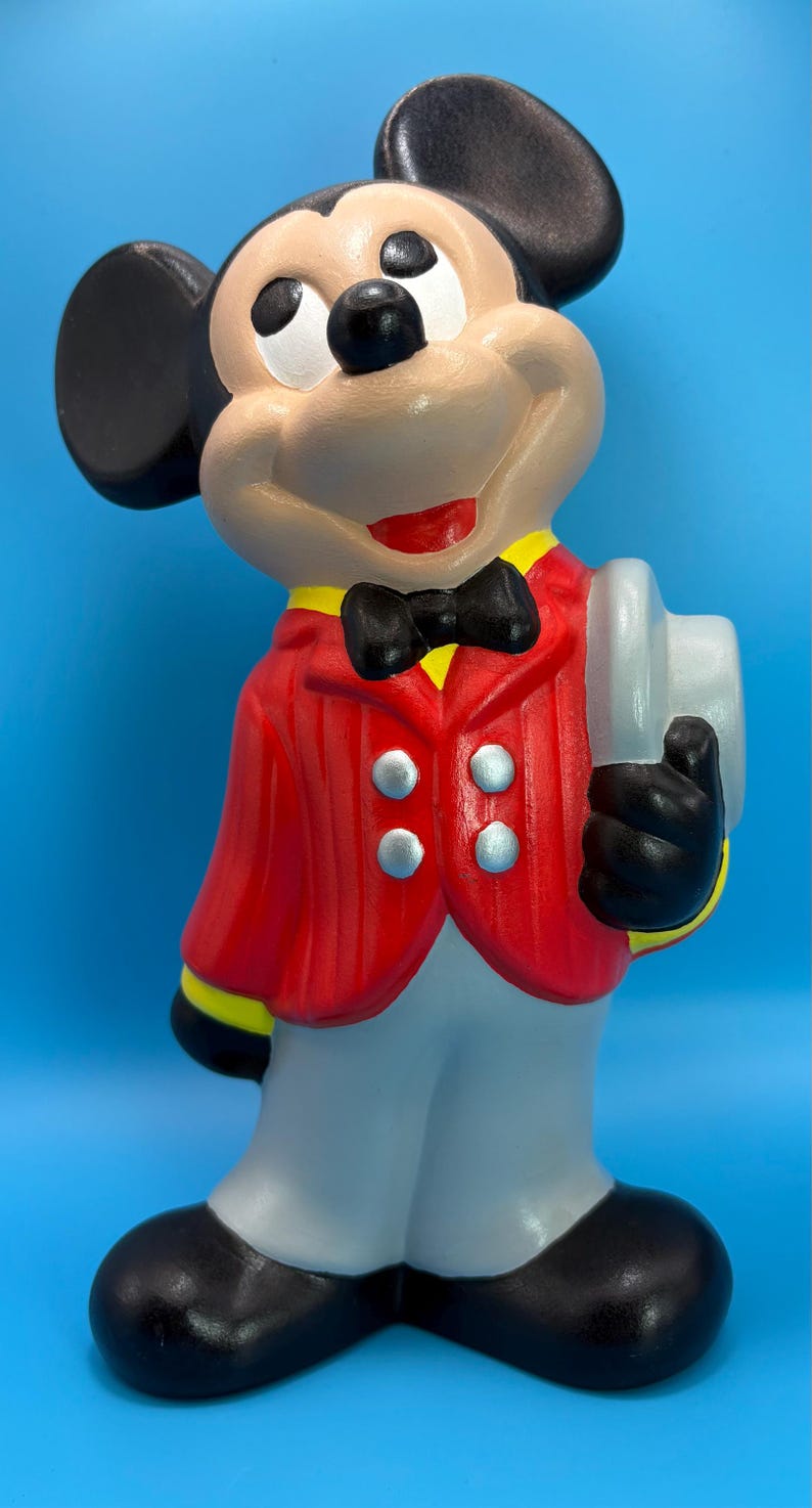 Peut inclure: Figurine en c&eacute;ramique de Mickey Mouse, v&ecirc;tu d'une veste rouge, d'un pantalon gris et de chaussures noires. La figurine porte un n&oelig;ud papillon noir, des oreilles noires et tient un objet blanc. Le fond est bleu uni.