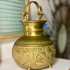 May include: A brass vessel with a rounded body, a flared neck, and a curved handle. The exterior features intricate embossed floral patterns and a textured band around the shoulder. The vessel stands on three small, short, cylindrical feet.