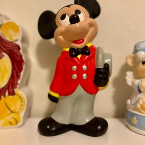 Peut inclure: Figurine en c&eacute;ramique de Mickey Mouse portant une veste rouge avec des d&eacute;tails noirs, un pantalon gris et des chaussures noires. La figurine tient un chapeau gris et porte un n&oelig;ud papillon noir. Fait partie d'une collection de figurines.