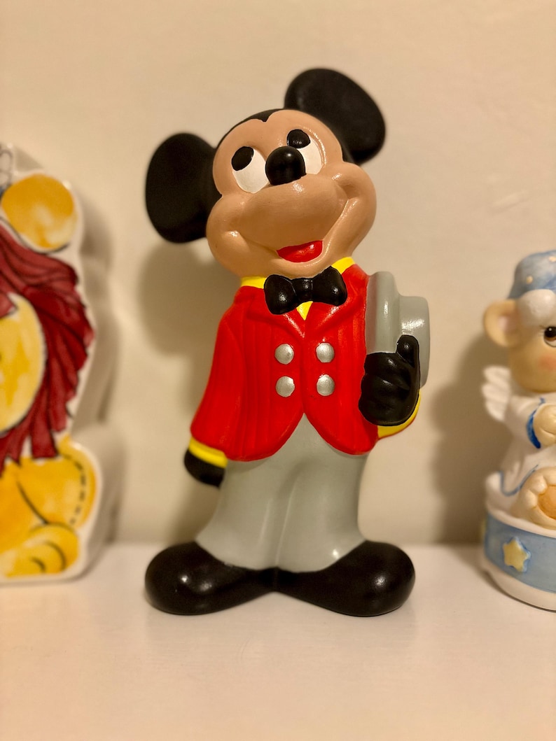 Peut inclure: Figurine en c&eacute;ramique de Mickey Mouse portant une veste rouge avec des d&eacute;tails noirs, un pantalon gris et des chaussures noires. La figurine tient un chapeau gris et porte un n&oelig;ud papillon noir. Fait partie d'une collection de figurines.