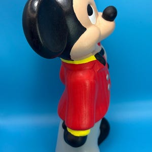 Peut inclure: Figurine en c&eacute;ramique de Mickey Mouse, avec une veste rouge &agrave; bordure jaune et un pantalon gris. Le personnage embl&eacute;matique a des oreilles noires, un nez noir et une expression souriante. Le fond est bleu uni.