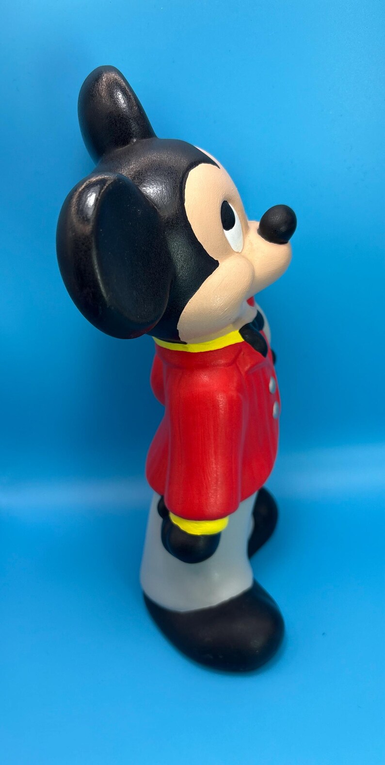 Peut inclure: Figurine en c&eacute;ramique de Mickey Mouse, avec une veste rouge &agrave; bordure jaune et un pantalon gris. Le personnage embl&eacute;matique a des oreilles noires, un nez noir et une expression souriante. Le fond est bleu uni.