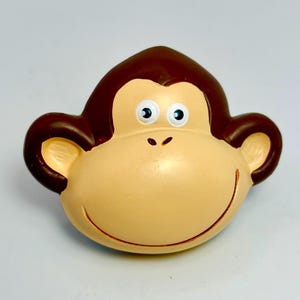 May include: A smiling monkey face figurine with a light brown face, dark brown hair, and large, round ears. The monkey has blue and white eyes and a small, curved mouth. The figurine is likely made of ceramic or a similar material.