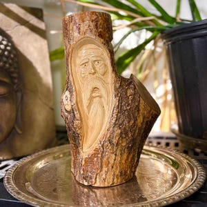 May include: A carved wooden sculpture of an elderly man's face, featuring a long beard and a furrowed brow, is carved into a section of tree trunk. The sculpture is displayed on a silver-coloured decorative tray. The wood has a natural, textured bark.