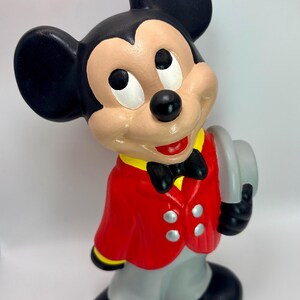 Peut inclure: Figurine en c&eacute;ramique de Mickey Mouse v&ecirc;tu d'une veste rouge avec des bordures jaunes, d'un pantalon gris et de chaussures noires. La figurine tient un chapeau gris et porte un n&oelig;ud papillon noir. Mickey a des oreilles noires, un visage beige et un nez noir.