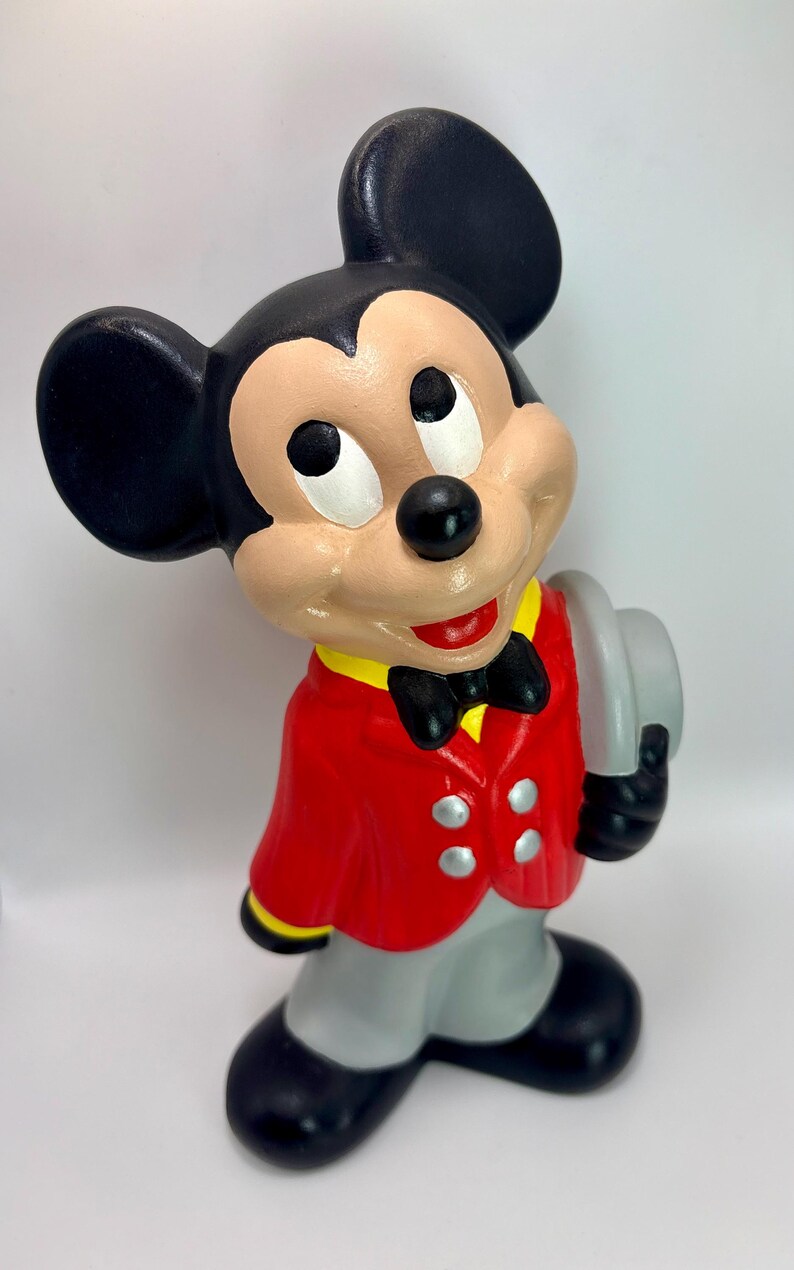 Peut inclure: Figurine en c&eacute;ramique de Mickey Mouse v&ecirc;tu d'une veste rouge avec des bordures jaunes, d'un pantalon gris et de chaussures noires. La figurine tient un chapeau gris et porte un n&oelig;ud papillon noir. Mickey a des oreilles noires, un visage beige et un nez noir.