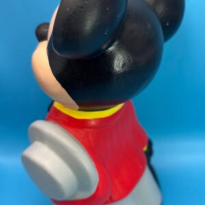 Peut inclure: Figurine en c&eacute;ramique d'un personnage de dessin anim&eacute; de souris. La souris a des oreilles et une t&ecirc;te noires, une veste rouge avec un col jaune, un pantalon gris et des gants blancs. La figurine est sur fond bleu.
