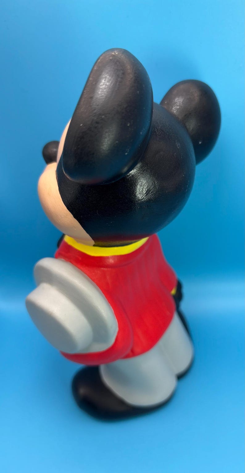 Peut inclure: Figurine en c&eacute;ramique d'un personnage de dessin anim&eacute; de souris. La souris a des oreilles et une t&ecirc;te noires, une veste rouge avec un col jaune, un pantalon gris et des gants blancs. La figurine est sur fond bleu.