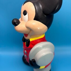 Peut inclure: Figurine en c&eacute;ramique de Mickey Mouse, peinte dans des couleurs classiques. La figurine porte une veste rouge avec des bordures jaunes, un short noir et un chapeau gris. La figurine a des oreilles noires, un visage beige et une expression joyeuse. Le fond est bleu uni.