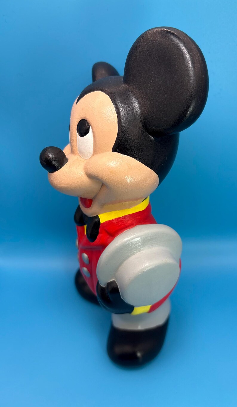 Peut inclure: Figurine en c&eacute;ramique de Mickey Mouse, peinte dans des couleurs classiques. La figurine porte une veste rouge avec des bordures jaunes, un short noir et un chapeau gris. La figurine a des oreilles noires, un visage beige et une expression joyeuse. Le fond est bleu uni.