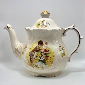 Op de afbeelding: Crèmekleurige keramische theepot met deksel en handvat, versierd met bloemmotieven en een centrale illustratie van een stel in een tuin. De theepot heeft gouden randen en een craquelé-afwerking.