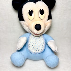 May include: A soft toy of Mickey Mouse in a seated pose. The doll has a black head and ears, a white face, and a blue body with a bib featuring a white and blue polka dot pattern. The doll's eyes are large and black.