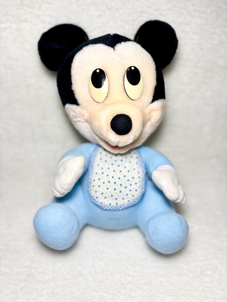 May include: A soft toy of Mickey Mouse in a seated pose. The doll has a black head and ears, a white face, and a blue body with a bib featuring a white and blue polka dot pattern. The doll's eyes are large and black.