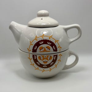 Puede incluir: Un juego de té individual de cerámica blanca. El juego incluye una tetera con tapa y una taza de té. La tetera y la taza de té están decoradas con un diseño floral marrón y amarillo. La tetera tiene un asa y un pico.