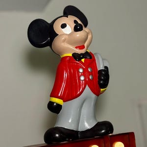 Peut inclure: Figurine de Mickey Mouse en veste rouge, pantalon gris et chaussures noires. La figurine a des oreilles noires, un visage bronz&eacute; et un sourire rouge. Il tient un objet gris et se tient sur une base rouge avec des lumi&egrave;res. Un objet de collection Disney classique.