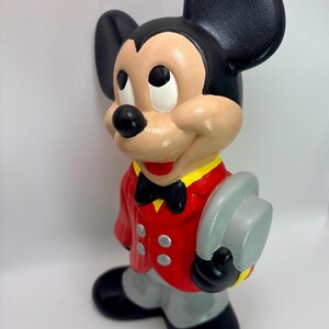 Peut inclure: Figurine en c&eacute;ramique de Mickey Mouse, v&ecirc;tu d'une veste rouge avec des d&eacute;tails jaunes, d'un pantalon gris et de chaussures noires. Mickey tient un chapeau gris et porte un n&oelig;ud papillon noir. Il a des oreilles noires et un visage beige.