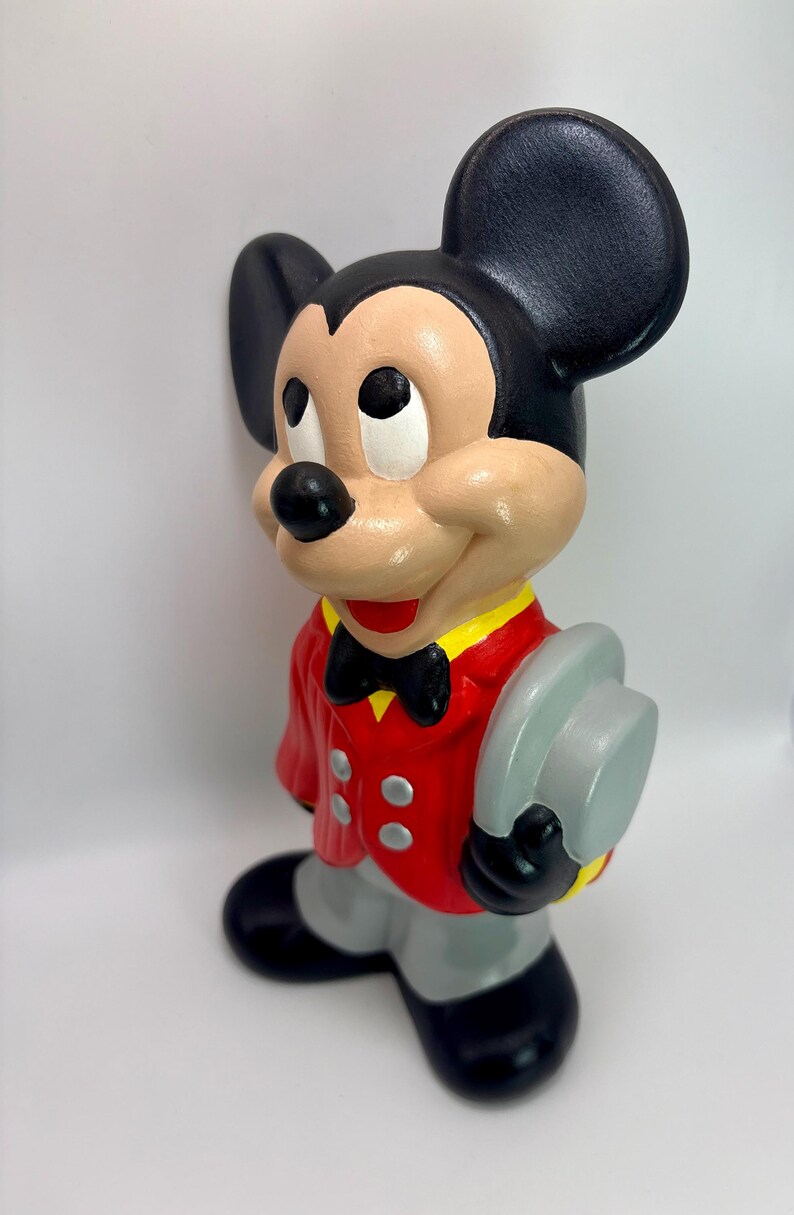 Peut inclure: Figurine en c&eacute;ramique de Mickey Mouse, v&ecirc;tu d'une veste rouge avec des d&eacute;tails jaunes, d'un pantalon gris et de chaussures noires. Mickey tient un chapeau gris et porte un n&oelig;ud papillon noir. Il a des oreilles noires et un visage beige.