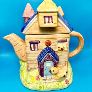 Può includere: Una teiera in ceramica stravagante a forma di casa, con esterno beige e dettagli blu e gialli. La teiera ha un manico, un beccuccio e un coperchio con un camino. Gli elementi decorativi includono un'anatra e fiori.