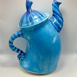 以下が含まれることがあります： 青色の陶器のティーポット。本体は明るい青色で、蓋、取っ手、注ぎ口には濃い青色のストライプ模様があります。取っ手はカールした端があります。ティーポットは光沢のある仕上がりで、白い背景に置かれています。