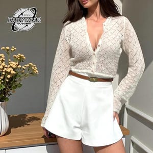 May include: A long-sleeved white lace cardigan with a button-down front and a V-neckline, paired with solid white high-waisted shorts and a thin brown leather belt with a gold buckle. The image includes a logo reading DAILY WEAR BY JEANINA.