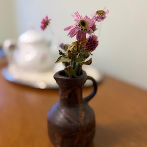 May include: A small, brown ceramic vase with a handle, holding dried pink and purple flowers. The vase has a textured surface with a leaf-like design. A white teapot is blurred in the background.