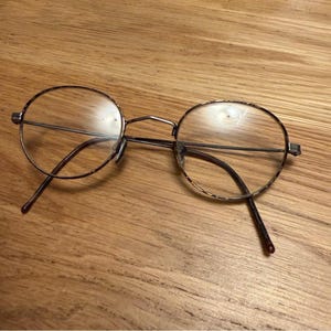 Peut inclure: Lunettes rondes avec une fine monture en métal doré. Les verres sont clairs et les branches sont marron foncé. Les lunettes reposent sur une surface en bois.