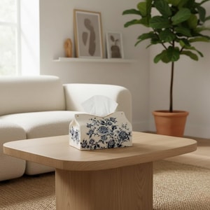 May include: A rectangular tissue box with a blue and white floral pattern rests on a light wood coffee table. The box contains white tissues and has a scalloped top. The room has a neutral colour palette.