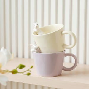 May include: Two stacked ceramic mugs, one cream and one lilac, each with a white bunny figurine perched on the rim. The mugs have rounded handles and are set against a white-striped background.