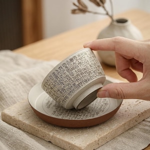 Puede incluir: Un juego de taza y platillo de cerámica blanca con borde marrón. La taza está cubierta de texto negro. El juego está sobre una superficie de piedra.