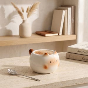 Op de afbeelding: Een witte keramische katvormige mok met een bruin handvat en gezichtsdetails, gevuld met cappuccino. Een zilveren lepel ligt op de tafel naast de mok.