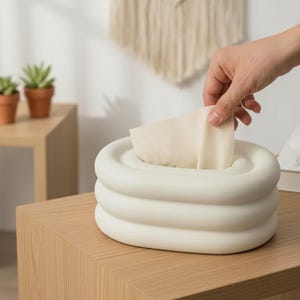 May include: A cream-coloured tissue box with a layered, rounded design. A hand is reaching in to take a tissue. The tissue box is on a light brown wooden surface.