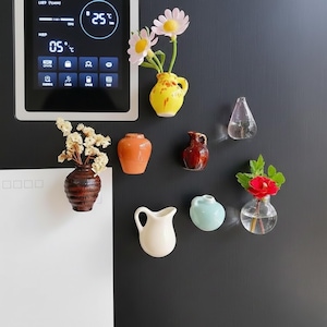 Może przedstawiać: Kolekcja małych, kolorowych ceramicznych wazonów i szklanych pojemników, z których każdy zawiera kwiaty lub zieleń. Wazony mają różne kształty i kolory, w tym żółty, pomarańczowy, brązowy i niebieski. Układ jest wyświetlany na ciemnej powierzchni.