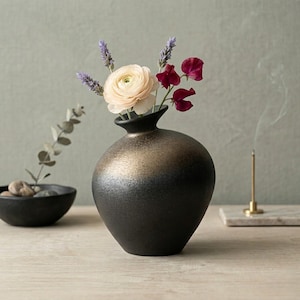 May include: A black ceramic vase with a gold gradient holds a bouquet of flowers. The arrangement includes a light pink rose, purple lavender, and deep red sweet peas. A small black bowl with rocks and an incense stick are also visible.