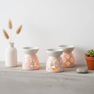 May include: Three pink ceramic oil warmers with white tops. One has the word 'LOVE' cut out, another a moon and stars, and the third flower cutouts. A small white vase with dried flowers and a potted succulent are also present, creating a serene display.