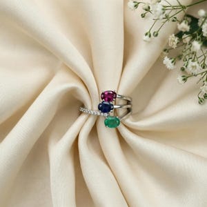 Op de afbeelding: Een zilveren ring met drie ovale edelstenen: een robijn, een saffier en een smaragd. De ring heeft een split-schachtontwerp met een rij kleine diamanten. De edelstenen zijn verticaal geplaatst.
