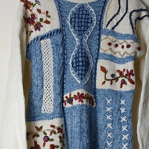 Könnte beinhalten: Ein Vintage-Pullover in Creme und Hellblau mit floralen Stickereien. Der Pullover hat ein Patchwork-Design mit Zopfstrick-, Lochmuster- und Kreuzstichdetails. Die Ärmel sind cremefarben.